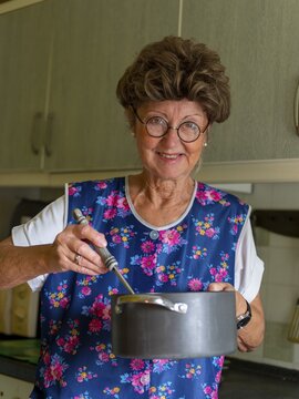 Granny with old smock apron, glasses and wig in the kitchen, holding cooking pot and ladle, Germany