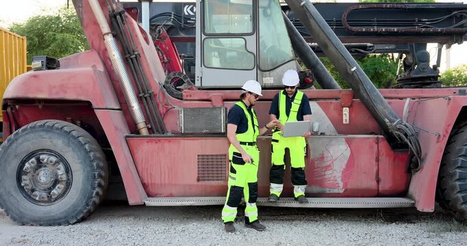 Adult female and male logistics team stand near reach stacker, using radio and laptop for cargo tracking, container handling safety checklist, freight yard coordination workflow onsite duty zone