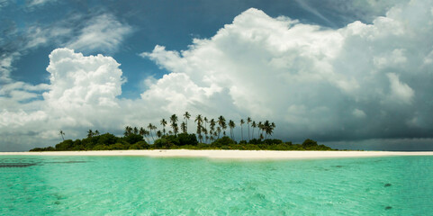 A scenic view of a tropical island with palm trees, a white sandy beach, and clear turquoise water under a bright cloudy sky in Maldives. Copy space available. © Aleksandr