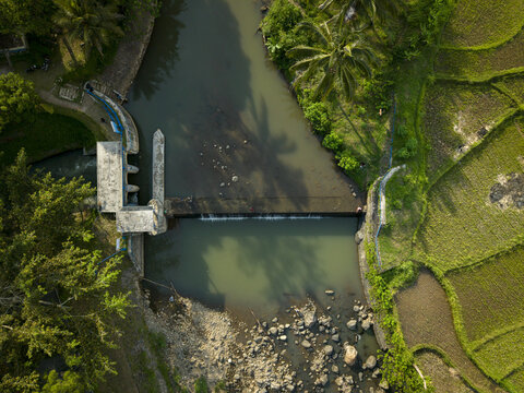Aerial view of Tangsi Water Gate on a river surrounded by lush green rice fields and palm trees in Kecamatan Slawi, Jawa Tengah, Indonesia.