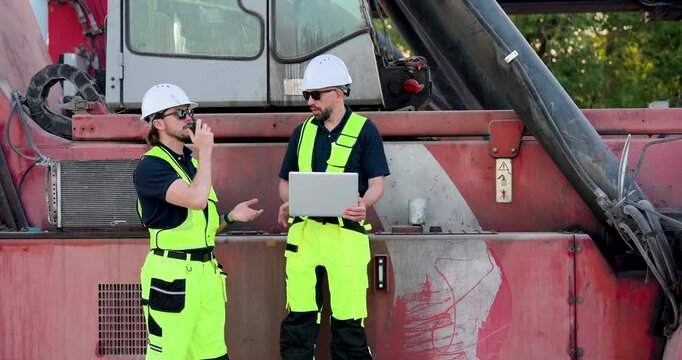 Adult Caucasian male logistics worker speaks by radio while colleague checks laptop, updating freight dispatch plan, container yard safety, cargo tracking, documentation workflow near reach stacker