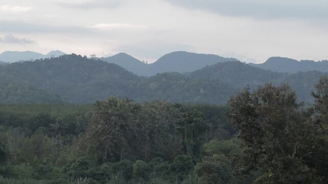 A beautiful panoramic view of layered tropical mountain ridges and lush green forests under an atmospheric overcast twilight sky.