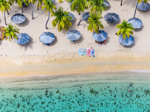 Aerial view of people sunbathing on a white sand beach with palm trees, blue umbrellas, and turquoise water in English Harbour, Saint Paul, Antigua and Barbuda.