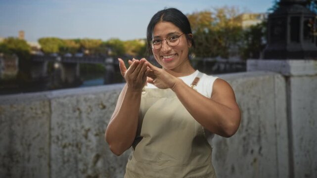 Woman in apron claps hands on street by stone bridge parapet while smiling and adjusting glasses; joy warmth friendliness.