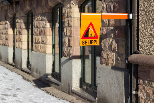Warning sign with text "SE UPP!" displayed on a building exterior, featuring stone walls and snow-covered ground in a city setting