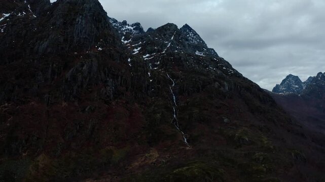 Mid-shot of steep mountain cliffs with snow patches near the peaks, as a river flows downward, revealed by a slight upward pan of the camera.