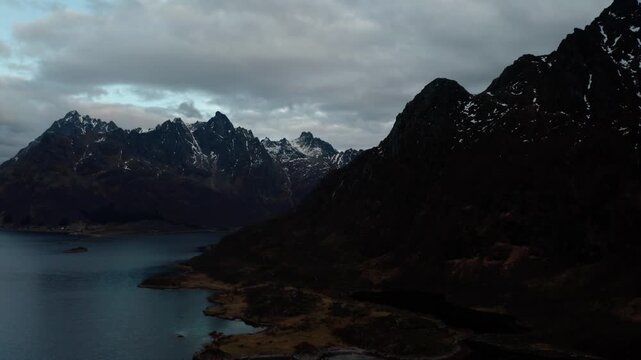 Wide shot of tall, pointy mountains surrounding a lake, with snow patches on rocky slopes under dark clouds, creating a moody and dramatic atmosphere.