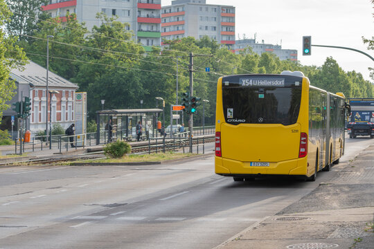 Germany, Berlin, June 1, 2026. A yellow Berlin bus marked X54 travels past a tram stop near residential apartment blocks. Linden and maple trees line the roadside in warm summer light.