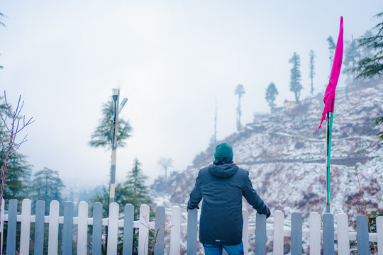 Person standing by a wooden fence overlooking a snowy mountain landscape in winter, Bhaderwah, Jammu, India