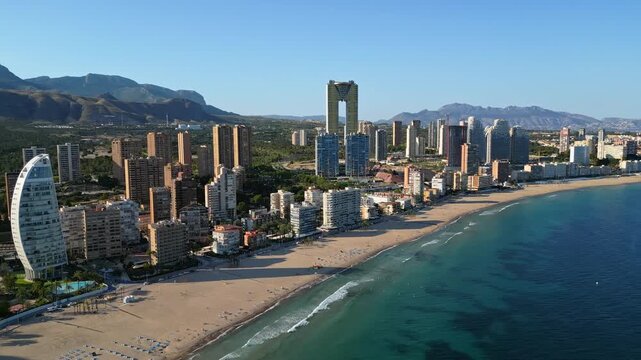 Aerial drone cinematic shot over sunny Benidorm beach showing modern skyscrapers and calm mediterranean sea waves.