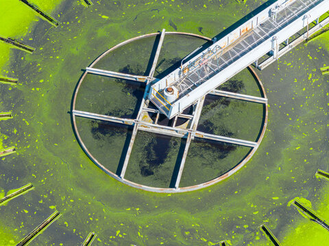 Aerial view of a circular wastewater treatment tank covered in bright green algae with a central rotating bridge in Prato, Tuscany, Italy.