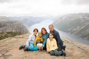 Fototapeta premium Family, enjoying the hike to Preikestolen, the Pulpit Rock in Lysebotn, Norway on a rainy day, toddler climbing with his pet dog the one of the most scenic fjords