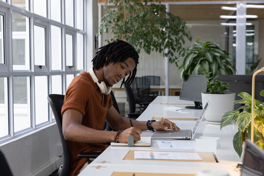 African American man writing in notebook in office with laptop, headphones, rust top, copy space