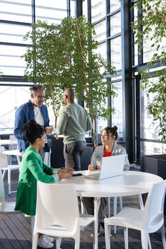 Diverse coworkers wearing business attire meeting around round table in office atrium with laptop