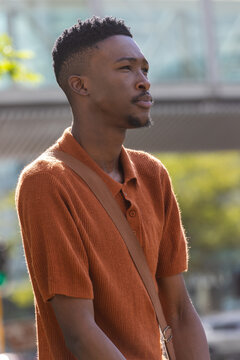 African man standing gazing right in sunny plaza wearing orange polo, crossbody strap, lip piercing