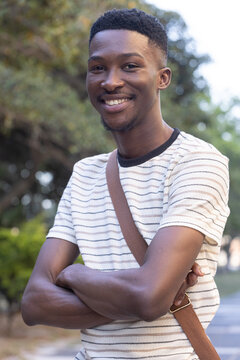 African American man standing on path smiling, wearing striped tee and brown crossbody strap