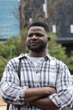 African American man standing arms crossed, looking left in plaza wearing plaid shirt and bag strap
