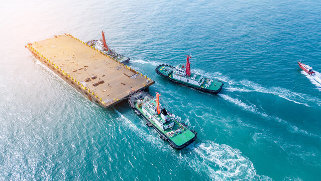 Tugboats towing a large empty flat deck barge through the turquoise ocean. Heavy marine logistics, offshore construction transport, and commercial maritime shipping operations.