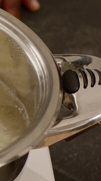 Vertical video: Steadying pan, hand gripping black knob as stove heating, checking pale liquid
