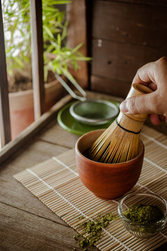 Close-up of a hand using a traditional bamboo whisk to mix green matcha powder in a ceramic bowl. Authentic Japanese tea ceremony preparation concept on a bamboo mat.