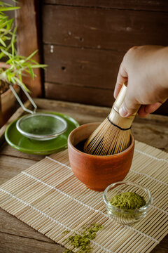 Close-up of a hand using a traditional bamboo whisk to mix green matcha powder in a ceramic bowl. Authentic Japanese tea ceremony preparation concept on a bamboo mat.