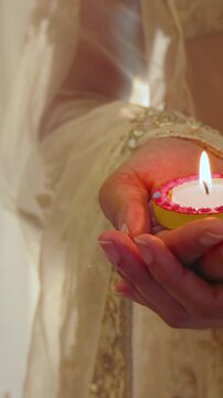 Vertical video: Flame drawing woman at home wearing cream sari, holding diya with pink rim for puja