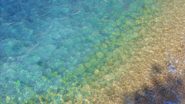 Crystal clear turquoise sea water ripple surface with colorful pebble stones under the shallow beach wave. Natural background texture of clean tropical ocean shore during summer holiday vacation.