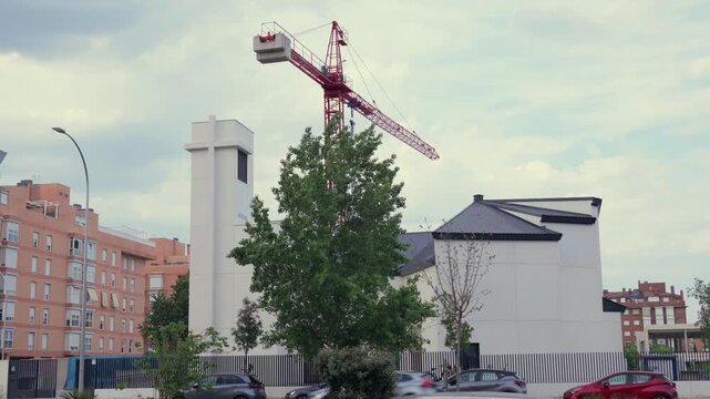 A daytime wide shot of a modern white Catholic parish church building under construction in Madrid, Spain. A large red tower crane stands behind the contemporary church structure and bell tower under 