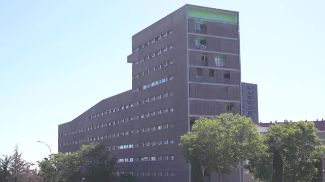A daytime wide shot of a contemporary multi-story residential apartment block with a dark facade and light green accents on the upper balconies in Madrid, Spain. In the foreground, several lush green 