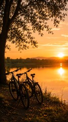 Fototapeta premium Two bicycles parked under a large tree by a serene lake, reflecting the vibrant orange and yellow hues of a beautiful sunset.
