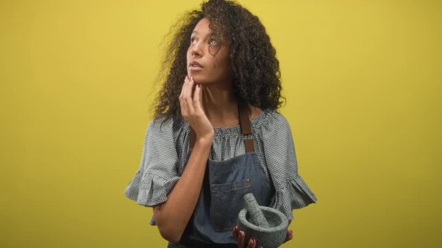 Teenage woman in apron holding mortar and pestle while hand touches cheek in studio; contemplation domestic craft.