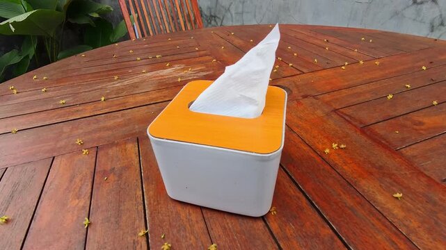 Video of tissue paper blowing in the wind inside a tissue box sitting on a warm wooden table. The paper napkin slightly protrudes from the top. Tiny yellow petals scatter around the tabletop. 4K Foota