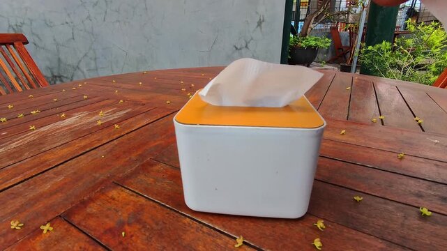 Vertical Footage of a hand picking up a tissue in a box. Tissue box with orange top and white edges. Tissue pulled out, slightly crumpled, mid-use. Modern, clean design sits on warm-toned planks. 4K V
