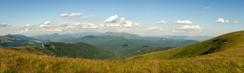 Panoramic view of undulating green hills, distant forested mountain ridges and clouds beneath clear blue sky