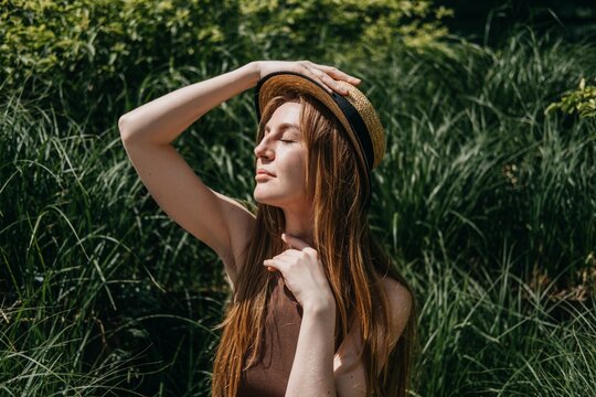 Woman sits on outdoor bench surrounded by green plants holding hand to straw hat. Managing urban heat stress, body hydration techniques , essential sun safe behaviors, summer fatigue prevention