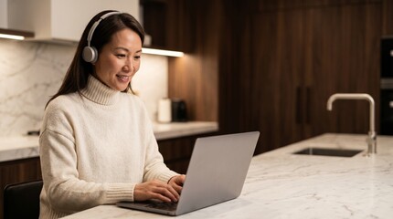 Fototapeta premium Smiling Asian woman working on laptop with headphones in modern kitchen.