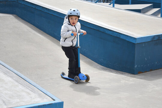 A young boy rides a blue threewheeled scooter at a concrete skatepark