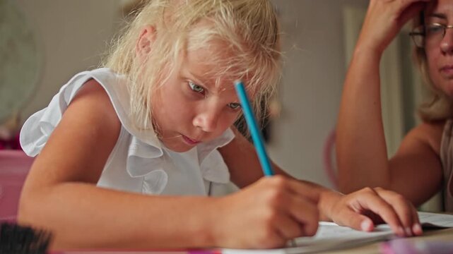 Schoolgirl writing homework in notebook at desk, mother nearby supervising. Home education and primary school learning