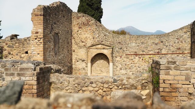Ancient Pompeii wall with a small shrine niche, rough stone masonry, brickwork, cypress tree, and Mount Vesuvius in the background.