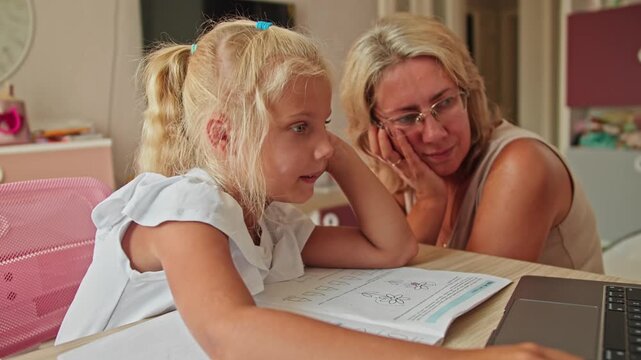 Schoolgirl and mother looking at laptop screen while doing homework together. Remote education and family learning environment