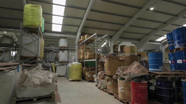 Large recycling warehouse stores bulk plastic waste bags, cardboard boxes and blue chemical drums on pallets