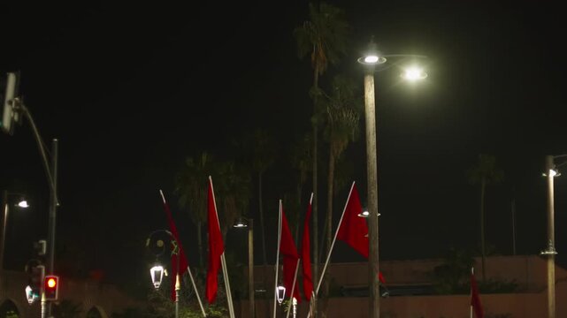 Rows of red Moroccan flags line an illuminated city boulevard at night with palm trees and street lamps