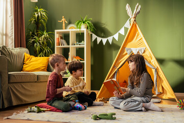 Girl reading a book to two boys in a playroom with teepee tent © MexChriss
