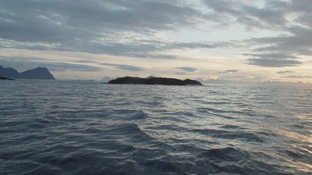 A vast ocean with small rocky islands in the foreground and distant mountains, under a mostly cloudy sky with subtle sunlight, creating a serene and cinematic seascape.
