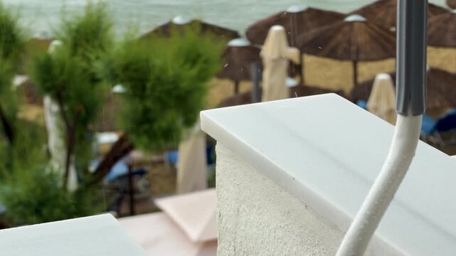 Summer rain falls onto a white stone balcony ledge at a beach resort, with blurred umbrellas, palm greenery, loungers and the sea in the background.