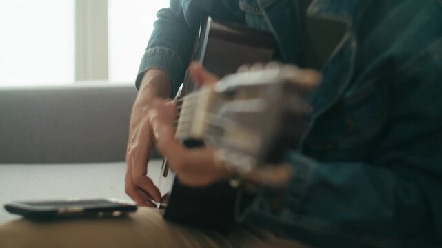 A close up shot of a man in a denim jacket playing a classic guitar while interacting with a smartphone app, showcasing authentic remote learning or digital nomad lifestyle.