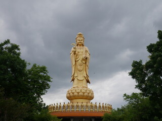 Fototapeta premium Large golden Guanyin statue standing on a lotus pedestal under dramatic storm clouds