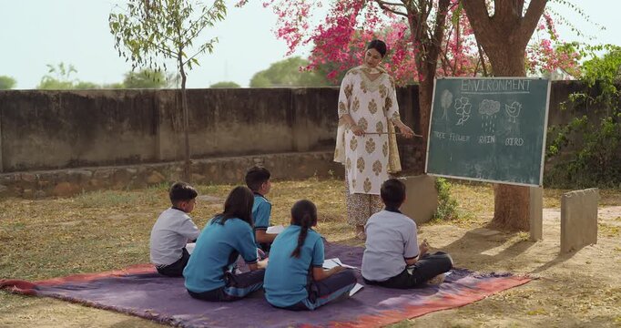 Indian young adult female hold stick pointing words talk speak teach to girl boy rural area place happy teen child group wear uniform sitting mat look board learn do follow tutor basic step at class