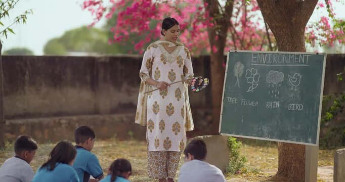 Indian happy young adult female standing hold color palette talk speak to girl boy group under tree rural area field tutor pointing stick on words teach look board read do study learn open class place