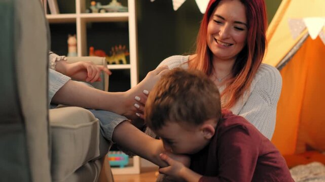 Happy mother and son tickling and smelling feet in playroom